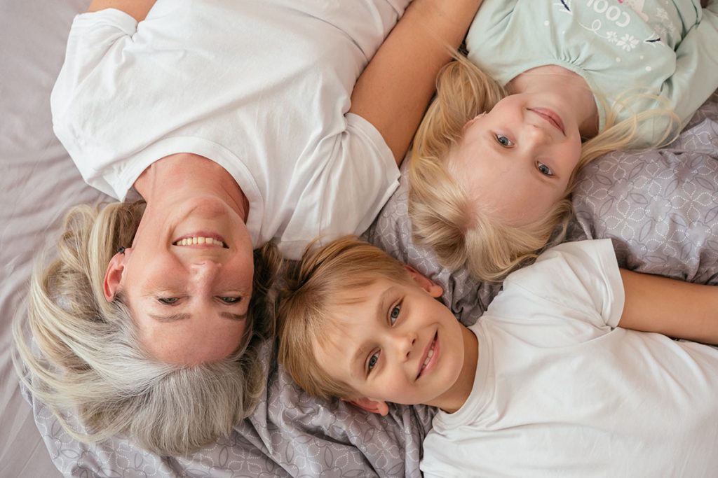 eine Mutter mit weißen Haaren und zwei Kinder von oben auf Bett fotografiert_alle mit weißem T-Shirt lachend und glücklich, gesund_ als Sinnbild für gesunde Schlafplätze, guten Schlaf, um Gesundheit von Lebewesen fördern zu können Unser Ziel ist Unterstützung bei Gesund Bauen - Gesund Wohnen - Gesund Schlafen.
