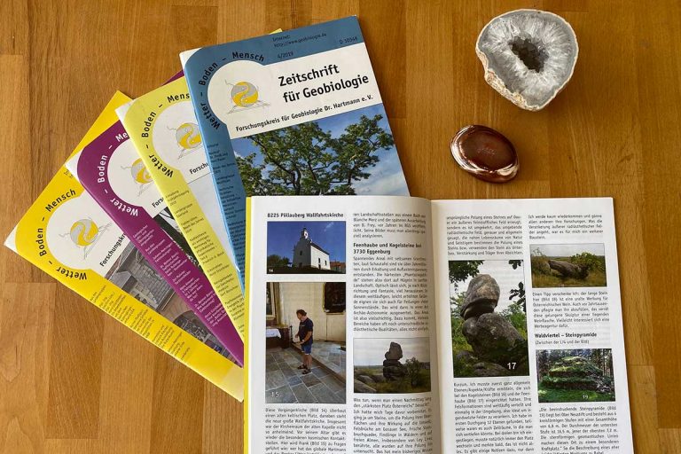 Aufgeschlagene Ausgabe der Zeitschrift WBM für Geobiologie, 3 weitere Ausgaben der Fachzeitschrift für Geobiologie im Hintergrund, eine Druse und Edelstein daneben.